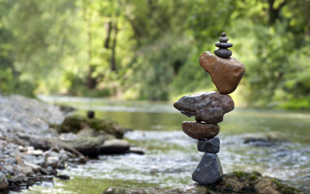 Image of several rocks balanced precariously on top of eachother set in front of a blurred body of water within a forest.