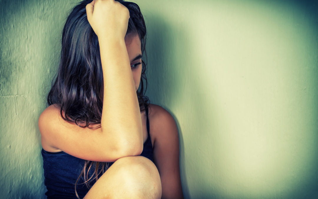 teenage girl sitting down with arm covering face and hand resting on head looking sad